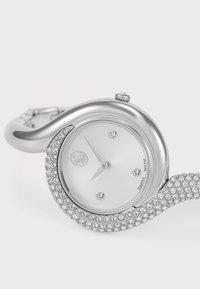 Silberne Uhr mit wellenförmigem Design, verziert mit funkelnden Kristallen. Weißes Zifferblatt, minimalistische Stundenmarkierungen und Gravur "Swiss Made".