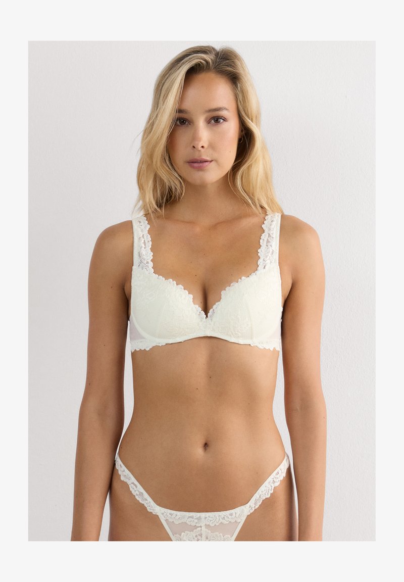 Vrouw met lang blond haar die een witte kanten beha en bijpassende kanten slipjes draagt, staand tegen een eenvoudige lichte achtergrond.
