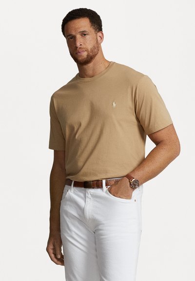 Beige bomulds t-shirt med rund hals og korte ærmer, der har et lille broderet logo på brystet, parret med hvide jeans og et brunt bælte.