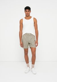 Un uomo che indossa una maglietta bianca senza maniche, pantaloni corti beige con motivo, calze rosa chiaro e sneaker bianche, in piedi davanti a uno sfondo bianco semplice.