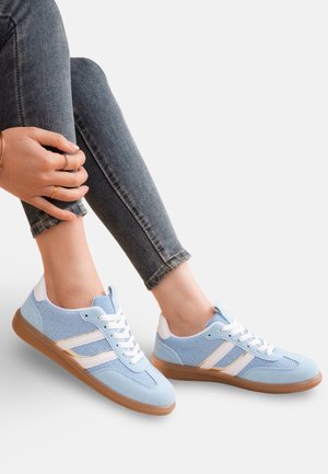 Personne portant des baskets bleu clair avec des rayures blanches et des semelles en gomme, associées à un jean skinny bleu foncé, assise jambes croisées.