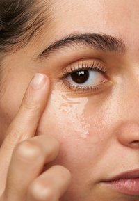Transparentes Augenserum wird mit dem Zeigefinger auf den Bereich unter dem Auge aufgetragen; die Haut erscheint glatt mit einem leichten Glanz. Ein braunes Auge ist sichtbar.