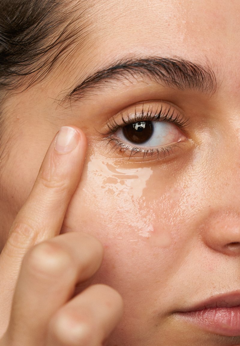 Transparentes Augenserum wird mit dem Zeigefinger auf den Bereich unter dem Auge aufgetragen; die Haut erscheint glatt mit einem leichten Glanz. Ein braunes Auge ist sichtbar.