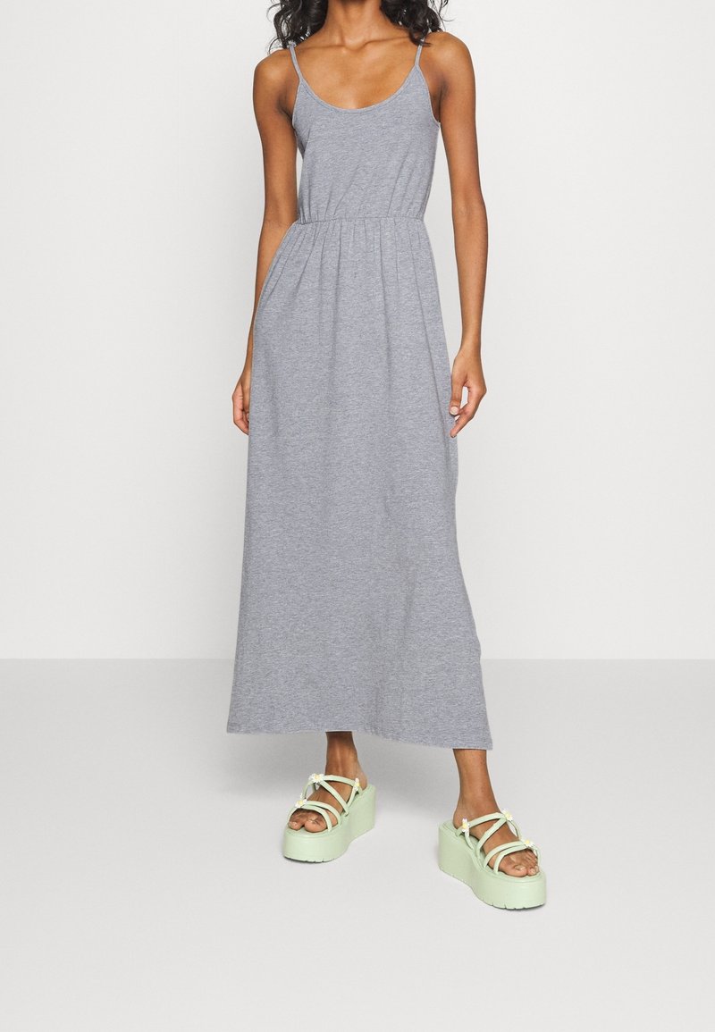 Graues Maxikleid mit dünnen Trägern, gerafftem Taillenbund und weichem Stoff. Kombiniert mit hellgrünen Plateausandalen mit floralen Akzenten.