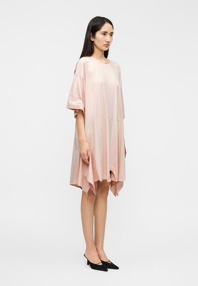 Φόρεμα με κοντά μανίκια σε απαλό ροζ ύφασμα, υπερμεγέθες, με ασύμμετρο τελείωμα  που είναι πιο μακρύ στο πίσω μέρος, λεία υφή, φορεμένο με μαύρα παπούτσια με τακούνι.