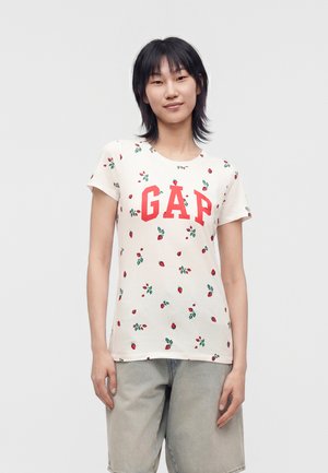 Ung kvinde med kort sort hår bærer hvid GAP t-shirt med rødt og grønt jordbærmønster og lysegrå shorts, står mod en hvid baggrund.