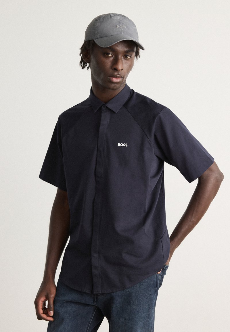 Camisa curta de manga azul-marinho com colarinho abotoado e logotipo "BOSS" bordado no peito. Combinada com jeans de denim azul-escuro e um boné cinza.