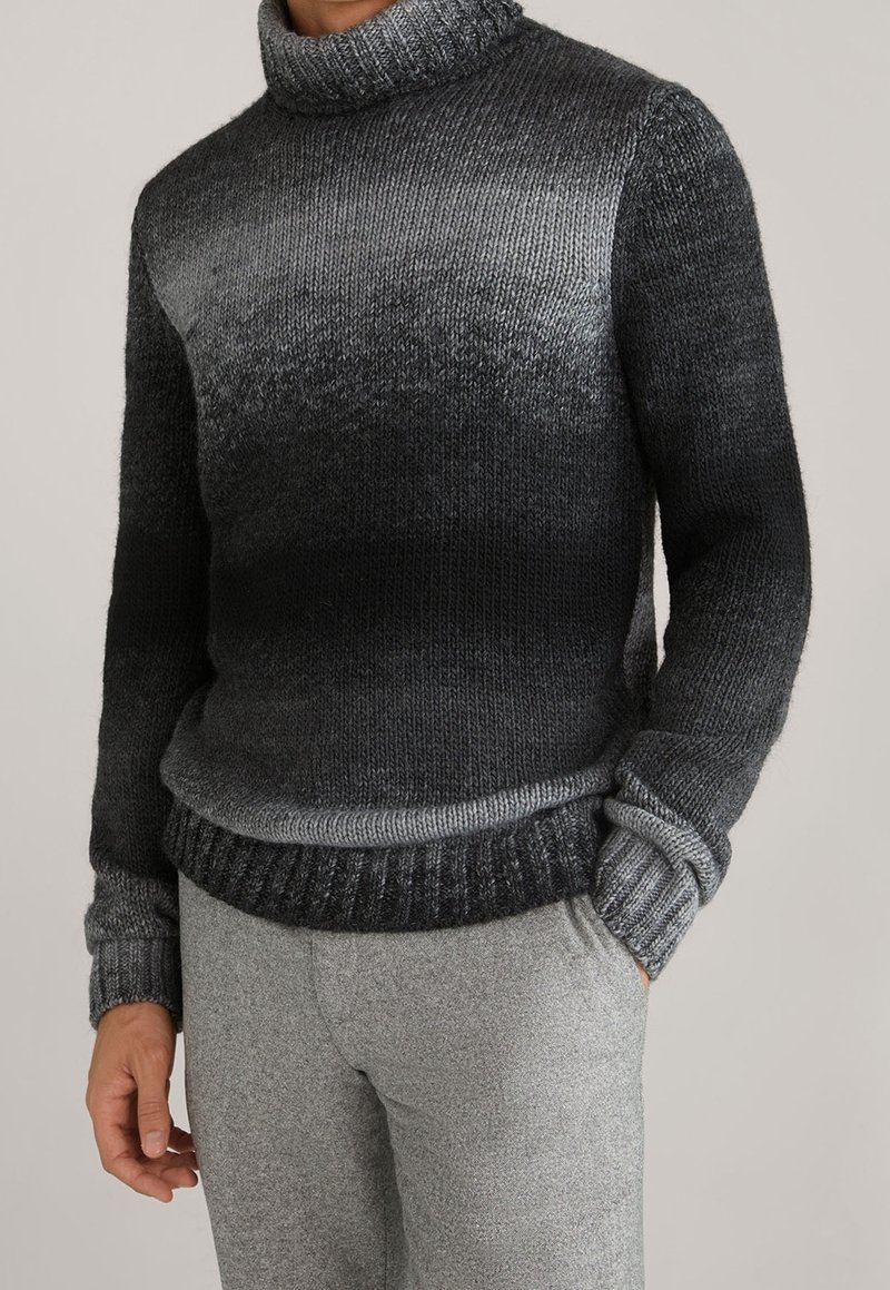 Homme portant un pull à col roulé en maille dégradé du gris foncé au noir et un pantalon gris clair, debout avec une main dans la poche.