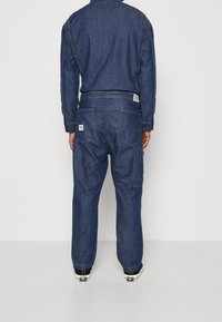 Combinaison en jean bleu foncé à coupe décontractée. Caractéristiques : deux poches arrière, coutures apparentes et une étiquette sur la ceinture.