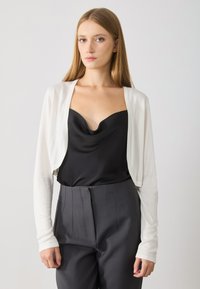 White cropped cardigan made of lightweight material over a black satin camisole, paired with dark high-waisted trousers. Smooth texture, fitted design.
