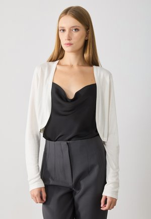 Cardigan corto bianco realizzato in materiale leggero sopra un top in satin nero, abbinato a pantaloni neri a vita alta. Texture liscia, design aderente.