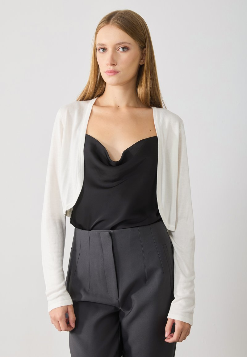 White cropped cardigan made of lightweight material over a black satin camisole, paired with dark high-waisted trousers. Smooth texture, fitted design.