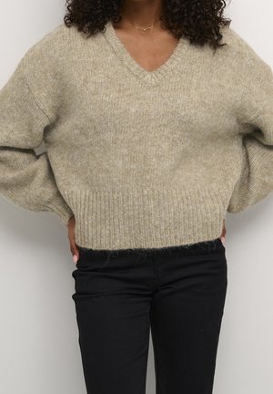 Personne portant un pull beige duveteux à col en V avec un ourlet côtelé et un pantalon noir, debout les mains posées sur les hanches.
