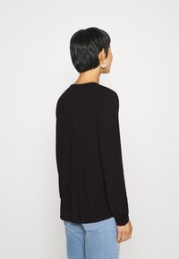 Svart blus med lång ärm och rund halsringning, med en plisserad detalj på baksidan och gatherings vid ärmarna, tillsammans med ljusblå jeans.