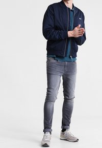 Blouson aviateur marine avec fermeture éclair, porté par-dessus un polo teal. Jean slim gris clair et baskets grises avec accents blancs.