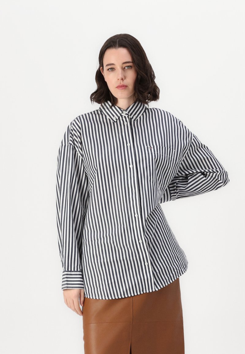 Camicia a righe con strisce verticali bianche e nere, colletto a punta e maniche lunghe. Presenta una tasca sul petto e una vestibilità rilassata.