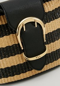 Svart läderrem med en guldbuckle-accent på en vävd väska med bruna och svarta ränder. Texturerade ytor framhäver kontrasten i designen.