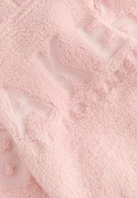 Pull en polaire en tissu rose doux avec une surface texturée et une inscription en relief dans un design tonal, créant une esthétique confortable.