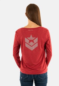 Rood langemouw T-shirt met een ontspannen halslijn. Heeft een ster- en chevronpatroon gemaakt van metalen studs op de achterkant.