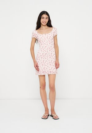 Femme debout et souriante, portant une robe rose à fleurs à manches courtes et des tongs blanches sur un fond blanc uni.