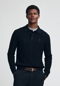 Svart stickad pullover med ett kabelmönster, med en krage och treknappslå, kombinerad med en grå t-shirt och mörka byxor.