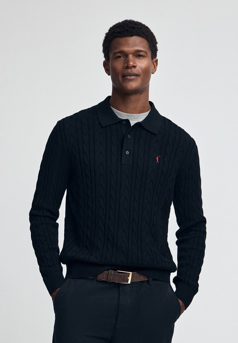 Svart stickad pullover med ett kabelmönster, med en krage och treknappslå, kombinerad med en grå t-shirt och mörka byxor.