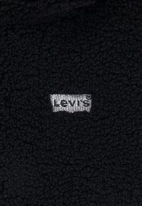 Crna flis tkanina s srebrnom oznakom Levi's ušivenom na materijalu, s teksturiranom, mekanom površinom.