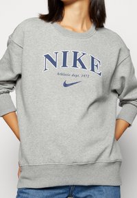 Grå sweatshirt med rundad halsringning, med en stor blå "NIKE"-logotyp, mindre text under, samt ribbade mudd och nederkant. Mjukt material.