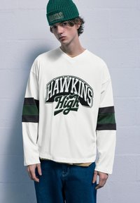 Biele tričko s dlhým rukávom s potlačou "HAWKINS High", so zelenými a čiernymi pruhmi, kombinované s modrými džínsy a zelenou rebrovanou čiapkou.