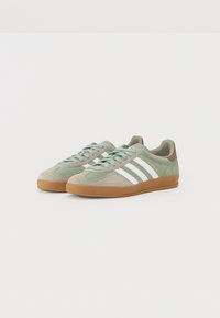 Ett par Adidas Gazelle-sneakers i ljusgrön och beige mocka med vita ränder och gummisulor på en vit bakgrund.