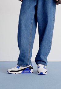 Sapatilhas brancas e pretas com detalhes azuis, apresentando uma unidade de amortecimento visível, combinadas com jeans de ganga azul de corte largo.