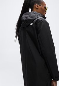 Μαύρο αδιάβροχο μπουφάν με κουκούλα, με διακριτή υφή και το λογότυπο The North Face στον ώμο. Μακριά, χαλαρή εφαρμογή.