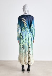 Langärmliges Kleid mit floralem Muster in Blau- und Grüntönen, mit einem taillierten Gürtel und aus einem glatten Stoff gefertigt. Schwarze High Heels Stiefel.