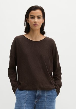 Jeune femme aux cheveux courts et foncés portant un haut ample à manches longues marron foncé et un jean bleu clair sur un fond blanc uni.