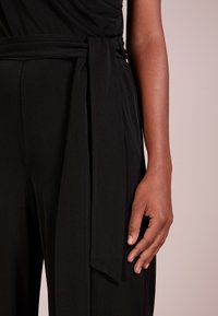 Svart, flödande jumpsuit med knutet midjeband, som har en mjuk tygstruktur och en åtsittande överdel, visad från midjan och uppåt.