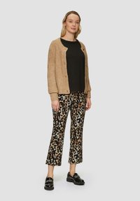 Beige strukturierte Strickjacke, schwarzer Pullover und weit geschnittene Hose mit Leopardenmuster. Schwarze Mokassins vervollständigen das Outfit. Neutraler Hintergrund.