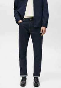 Mørkeblå denim jeans med lige snit, oprullede manchetter og synlig syning. Bæres med en lys skjorte og marineblå jakke, parret med sorte sko.