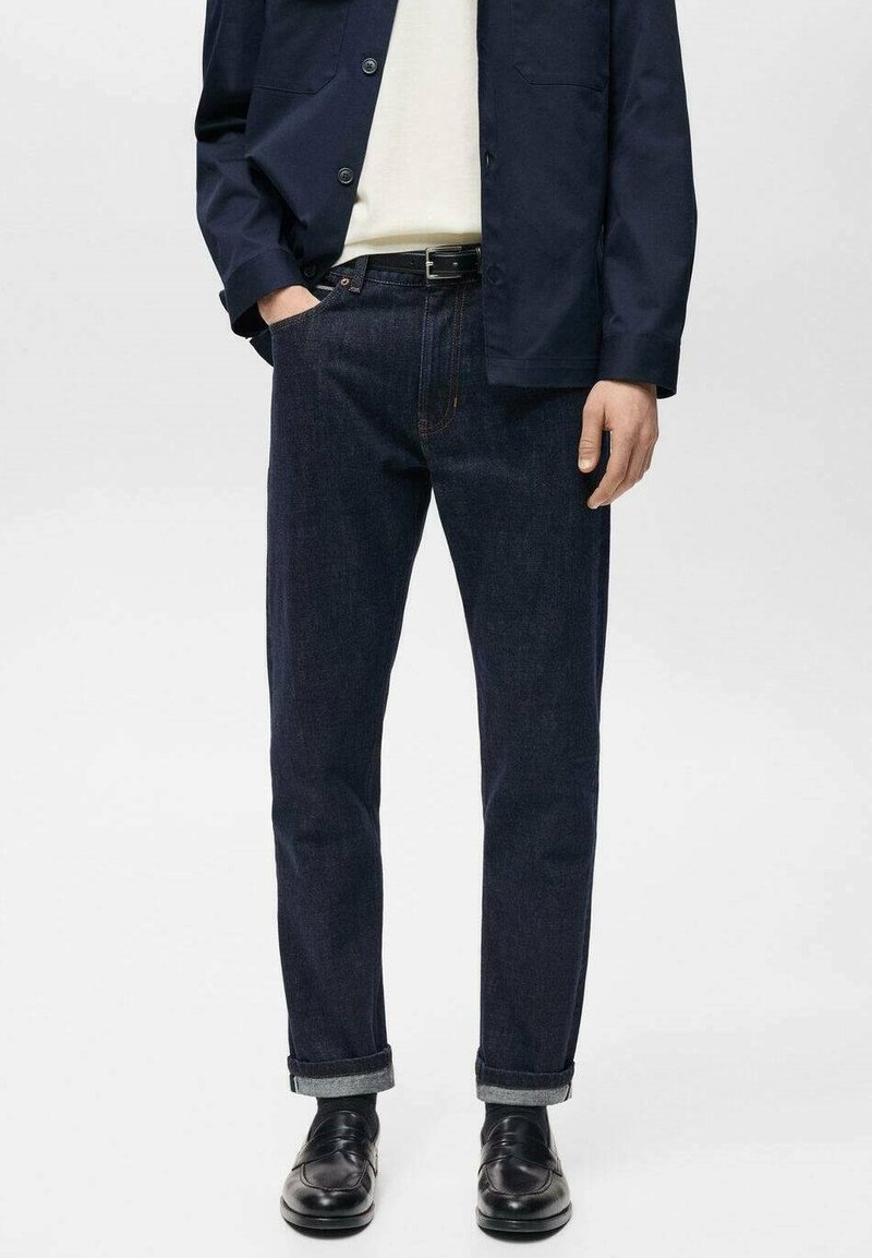 Mørkeblå denim jeans med lige snit, oprullede manchetter og synlig syning. Bæres med en lys skjorte og marineblå jakke, parret med sorte sko.