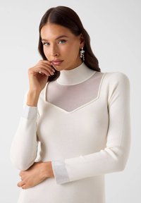 Robe blanche ajustée avec un col montant et un panneau transparent sur la poitrine, agrémentée de petites perles. Les manches longues présentent des poignets à motif métallique.