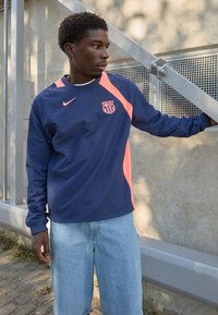 Langærmet marineblå og koralfarvet Nike tr�øje med Barcelona-logo, der har en rund halsudskæring og sporty design. Bæres med lyseblå shorts.