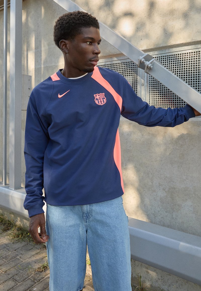 Long-sleeve navy and coral Nike jersey with Barcelona logo, featuring a round neckline and sporty design. Worn with light blue shorts.