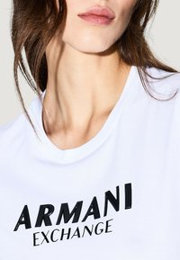 Camiseta blanca de algodón con cuello redondo y un logo impreso en negro "ARMANI EXCHANGE" con un detalle texturizado en las letras.