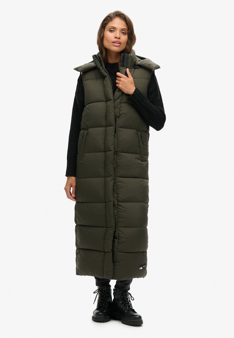 Langer, olivgrüner Steppwesten mit Kapuze, versehen mit horizontalen Steppmustern und zwei Seitentaschen. Getragen über einem schwarzen Pullover und einer Hose.