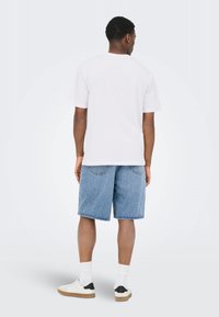 Mand stående med ryggen til iført en hvid t-shirt, blå denimshorts, hvide sokker og hvide sneakers med sorte hældetaljer.