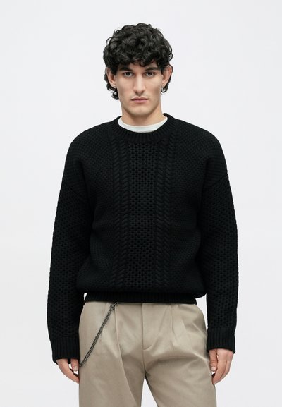 Νέος με σγουρά σκουρόχρωμα μαλλιά, φοράει μαύρο πουλόβερ με υφή, μπεζ παντελόνι και αλυσίδα στο αριστερό μέρος, στέκεται μπροστά σε ανοιχτό φόντο.