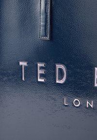 Τσάντα ώμου από μπλε ναυτικό δέρμα με υφή και λεία επιφάνεια, με ασημί ανάγλυφα γράμματα "TED BAKER" και "LONDON" στο μπροστινό μέρος.