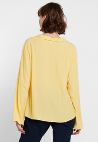 Femme aux cheveux bruns jusqu'aux épaules, portant un haut long aux manches amples de couleur jaune et un pantalon sombre, vue de dos sur un fond uni.