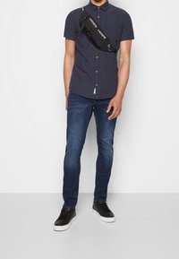 Camicia navy a maniche corte con colletto button-down, abbinata a jeans slim-fit blu scuro e sneakers nere con suola bianca. Borsa a tracolla nera.