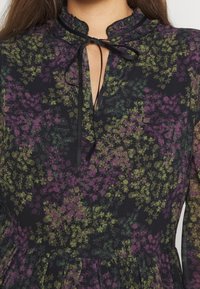 Svart blommig klänning med lila och gröna mönster, med en volangkrage, en nyckelhålsklippning fram och ett mjukt, lätt tyg.