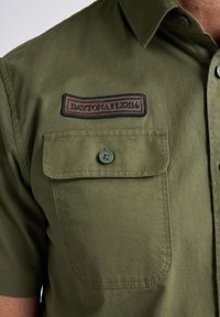 Camisa de manga corta verde oliva con un parche que dice "DAYTONA 152114", dos bolsillos en el pecho y un detalle de botones. Textura de tela suave.